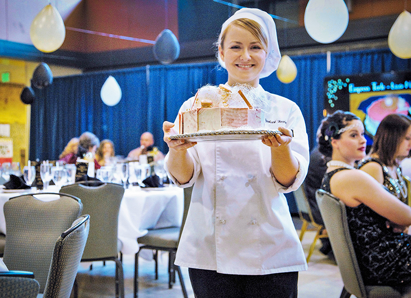 Lizzie Hartman stands in chef gear holding a cake